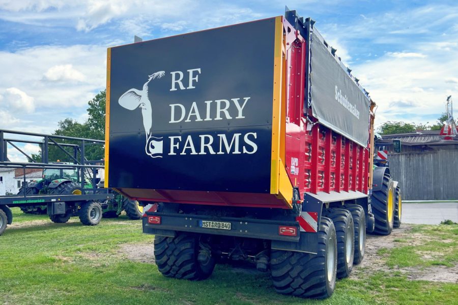 Ein Agraranhänger von hinten mit einen Schwarz-Weiß-Logo einer Kuh und dem Schriftzug „RF DAIRY FARMS“ steht auf einer Wiese. Daneben ist ein
