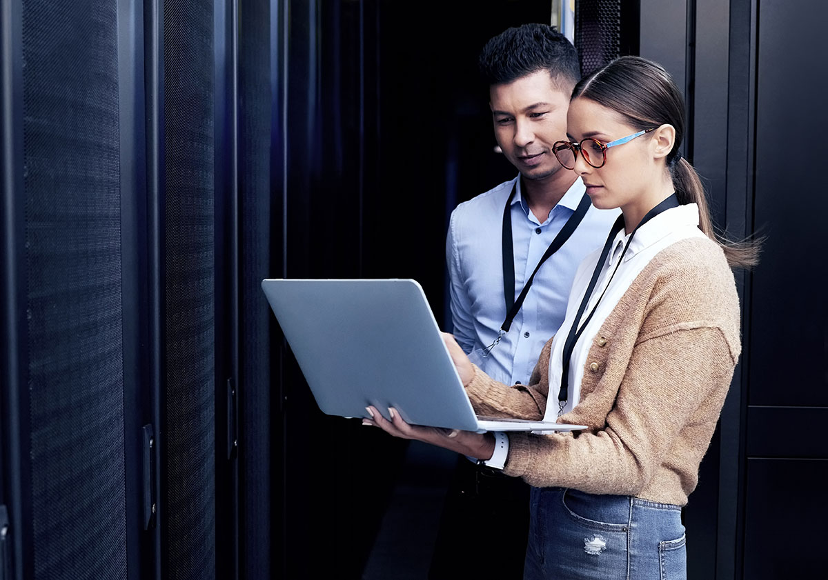 Eine Frau und ein Mann stehen vor einem Serverschrank. Die Frau hält einen aufgeklappten Laptop in der Hand und zeigt dem Mann etwas auf dem Bildschirm.